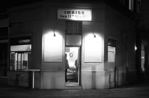 Nachts in einer Stadt: eine Eckkneipe, auf der "Imbiss von 11 bis 11" steht. Ein Mann steht im Türbereich.